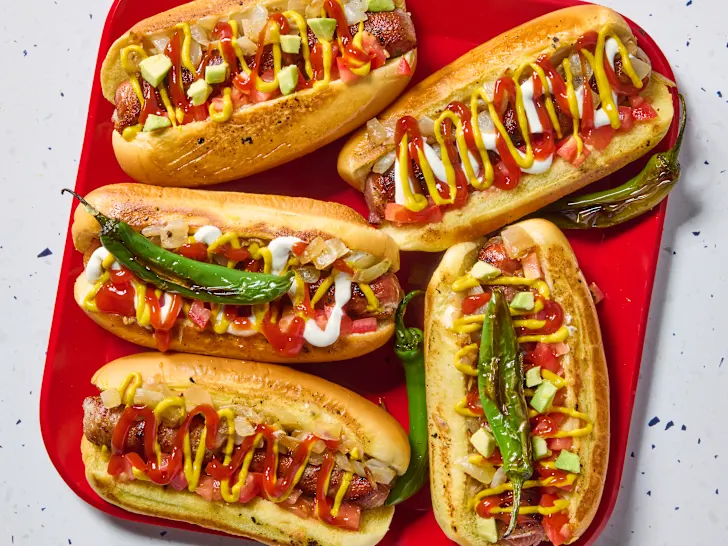The LA style hotdog shows grilled hotdog sitting atop a golden bun, the hotdog is wrapped in bacon, and topped with serano peppers, ketchup, mustard, and sour cream.